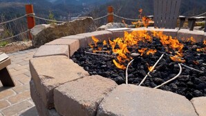 Boccia in pietra con tronchi ardenti, circondata da un muro in mattoni e una sedia Adirondack in legno.