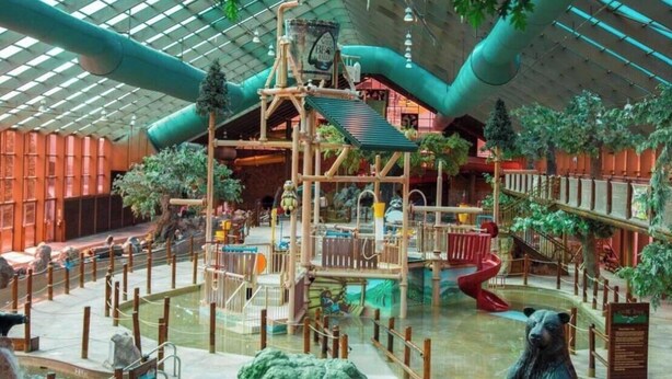 Un'area giochi interna con una struttura in legno per arrampicarsi, scivoli e una statua di un orso.