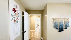 Un couloir doté d’un plancher en bois, une porte sur la gauche et un mur orné d’une œuvre d’art en macramé sur la droite.