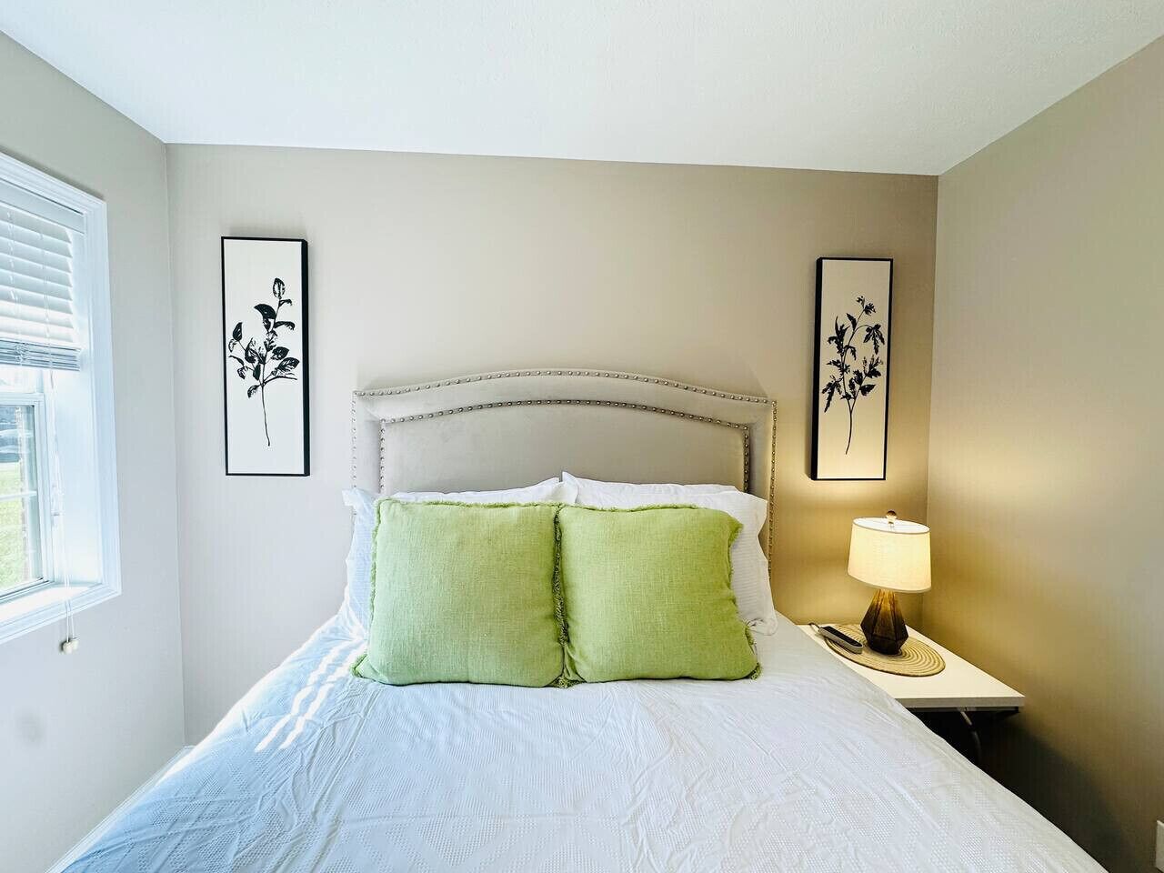 A neatly made bed with two green pillows, a bedside lamp, and two framed floral artworks on the wall.