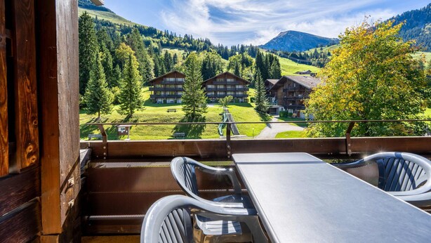 A wooden balcony with a table and chairs, overlooking a mountain landscape with buildings and trees.