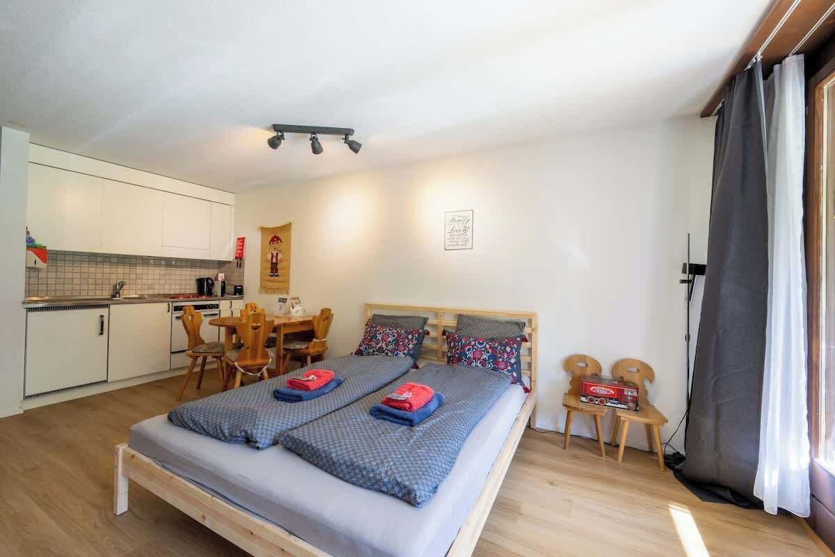 Una camera da letto con un letto, due comodini, una scrivania e una sedia. Si nota una piccola cucina in fondo.