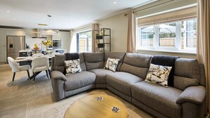A modern living room with a grey sectional sofa, a wooden coffee table, and a dining area with white chairs. - Host Stay The Sett (King's Lynn)