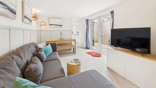 A modern living room with a grey sectional sofa, a wooden coffee table, a flat-screen TV on a white cabinet, and a dining area with a wooden table and bench. - Host Stay Surf Jewel (Saltburn-by-the-Sea)
