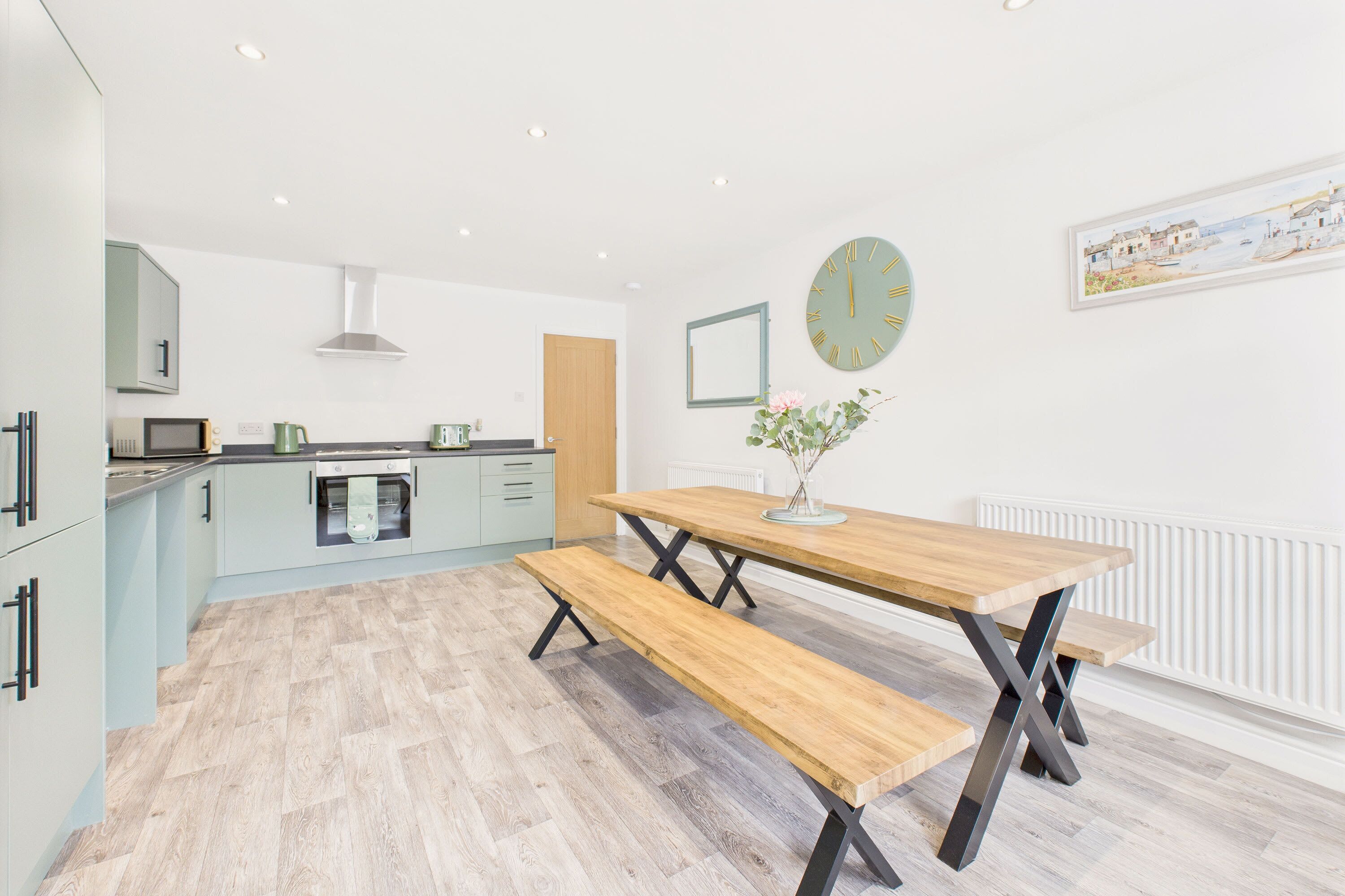 A modern kitchen with a wooden dining table and benches, a clock on the wall, and a radiator.