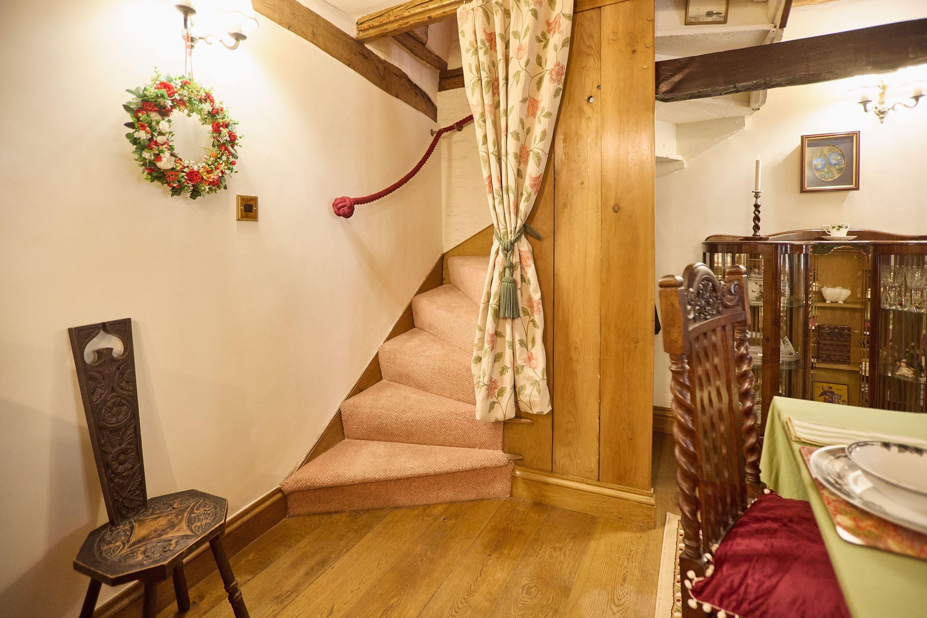 Un escalier orné d’une tenture à motifs floraux, une chaise en bois et une table recouverte d’une nappe verte.