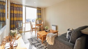 A modern living room with a sofa, a dining table, and chairs. There is a large window with curtains, a small table with a plant, and a radiator. - Host Stay 608 Hatton Garden (Liverpool)