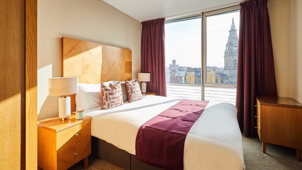 A hotel room with a large bed, wooden headboard, two bedside tables with lamps, a nightstand, and a view of a cityscape through large windows. - Host Stay 608 Hatton Garden (Liverpool)