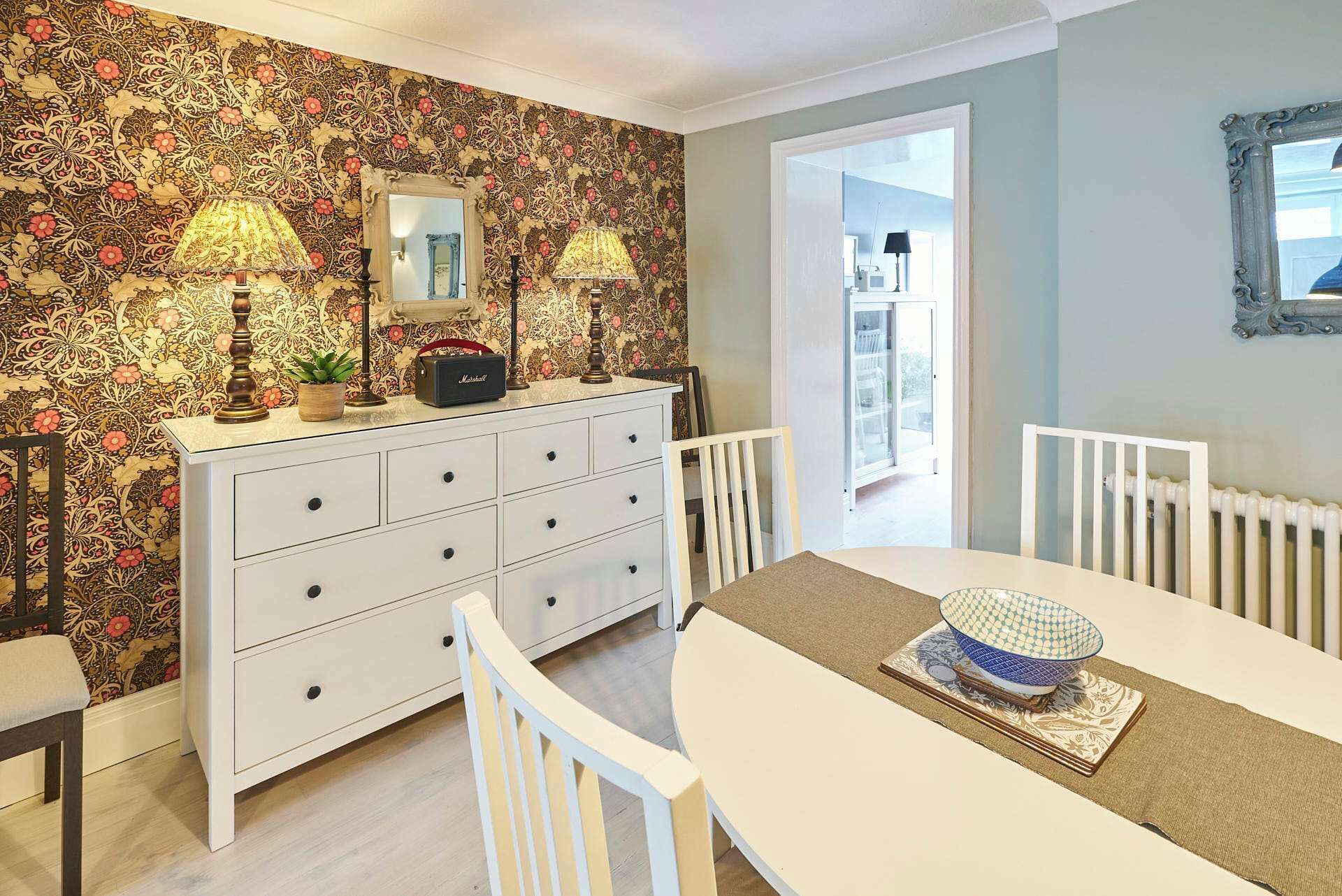 A dining area with a white table, chairs, and a patterned wallpaper. There is a buffet table with a bowl and a radio.
