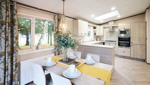 Et moderne køkken med indbyggede apparater, et spiseområde med et bord dækket til fire personer, og udsigt til naturen gennem store vinduer.