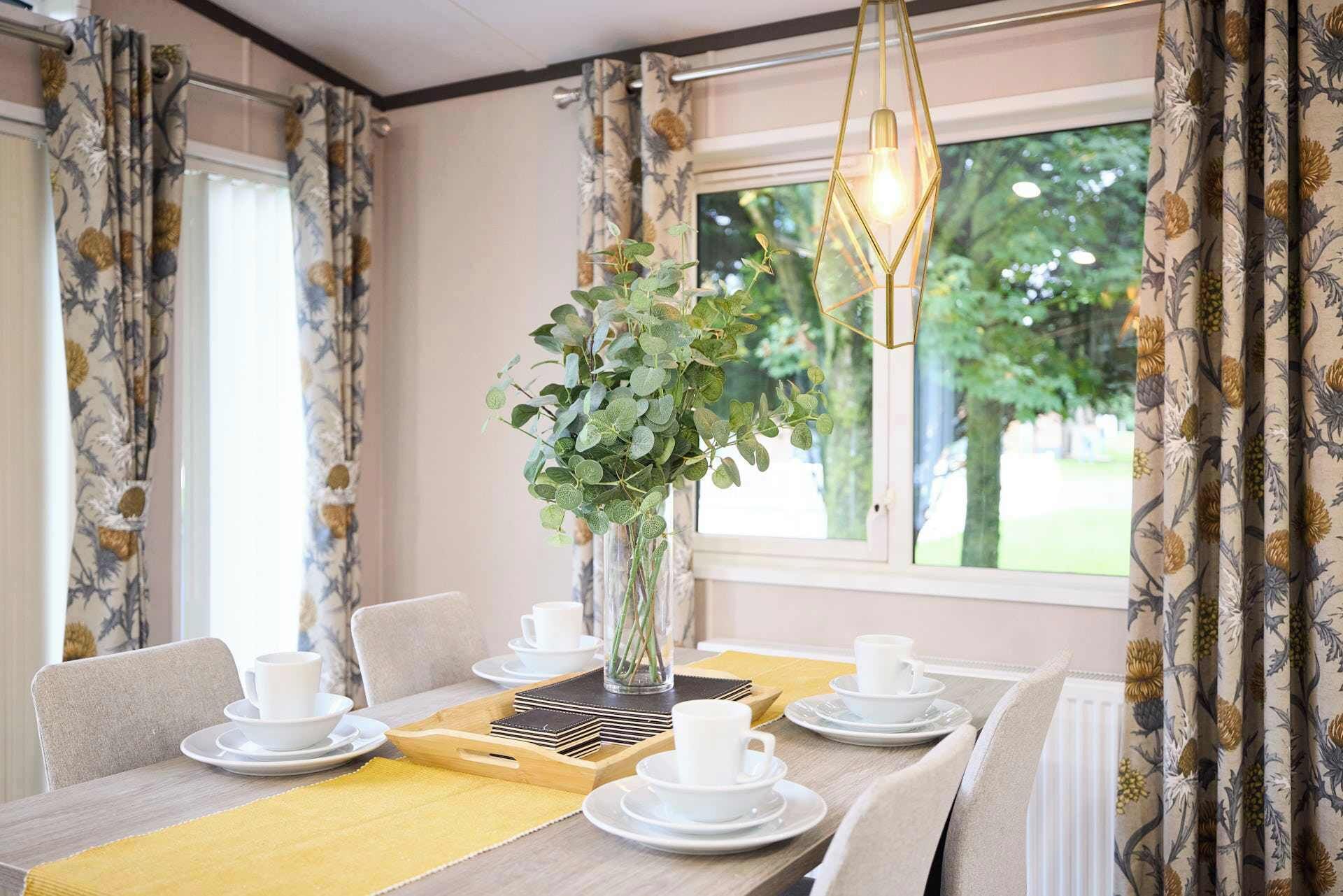 A dining area with a table set for four, a vase with green foliage, and a view of trees through the window.