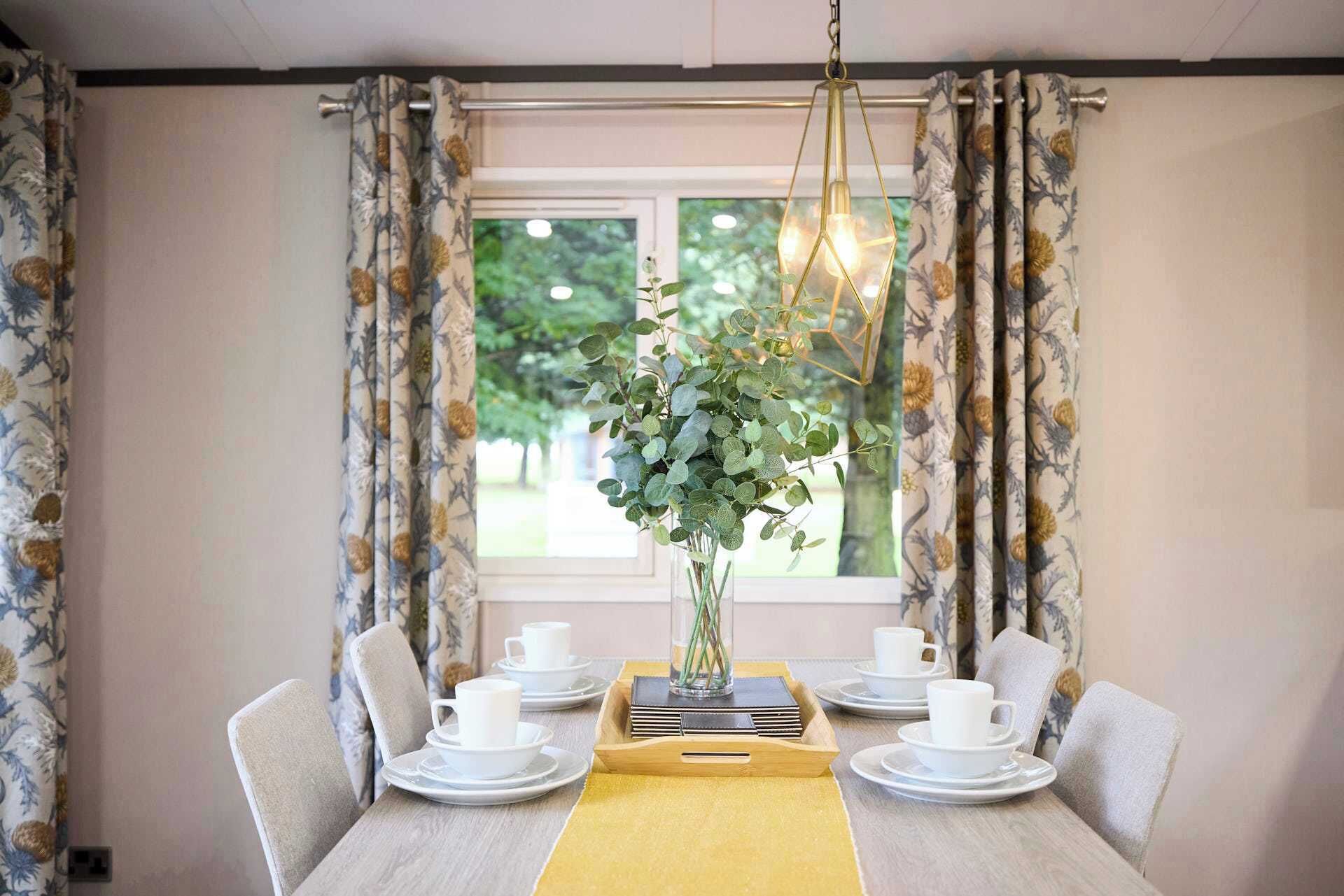 A dining area with a table set for four, a vase with greenery, and curtains with a floral pattern.
