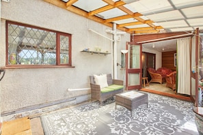 A covered patio area with a wicker sofa, a small table, and a patterned rug. - Host Stay Windles (Wotton-under-Edge)
