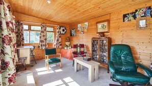 A cozy cabin interior with wooden walls, a green leather chair, a wooden table, and a window with floral curtains. - Host Stay Windles (Wotton-under-Edge)