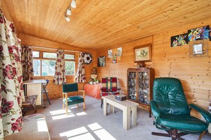 Interior acogedor de una cabaña con paredes de madera, un sillón de cuero verde, una mesa de madera y una ventana con cortinas florales.