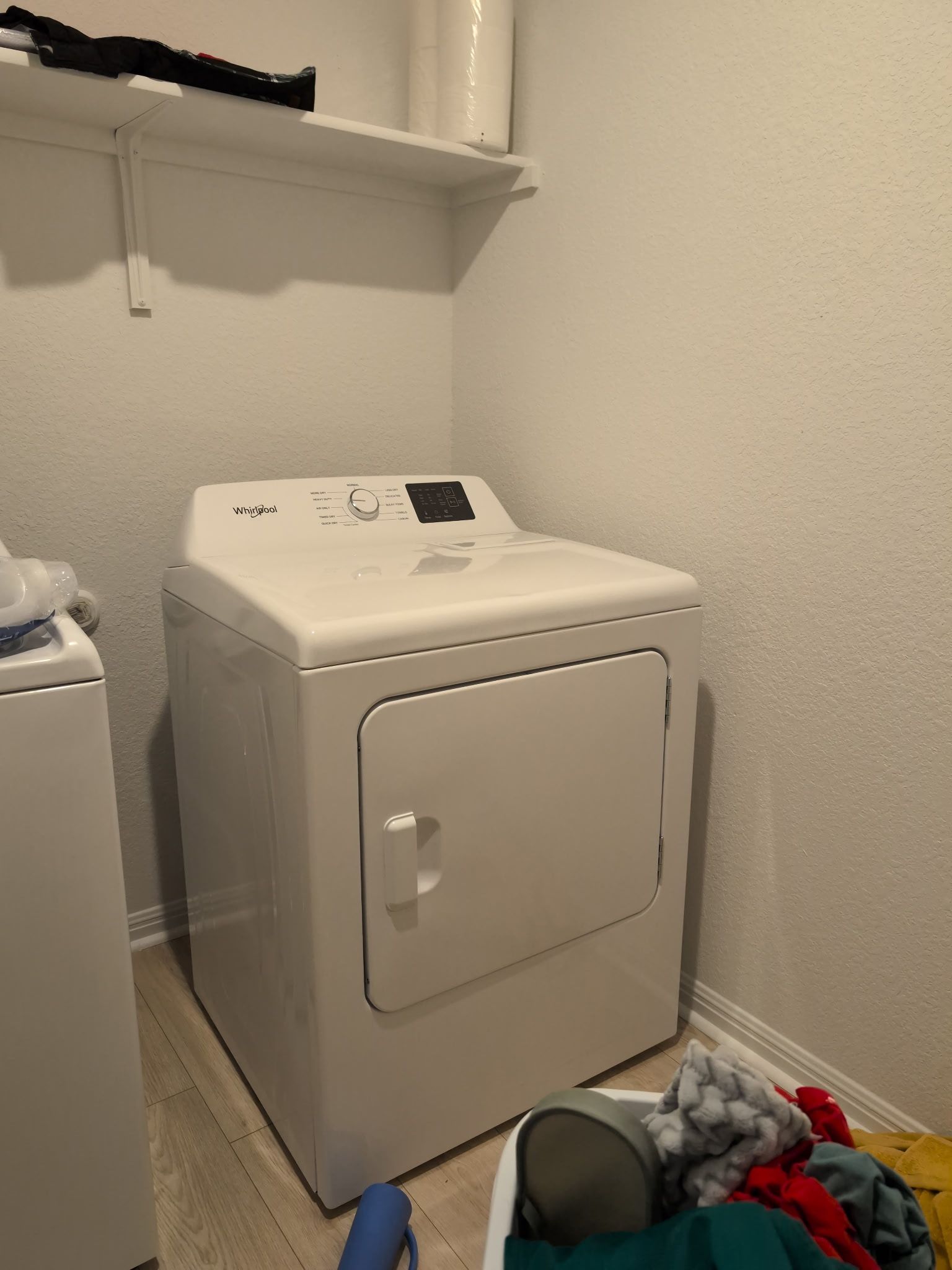 Una lavadora blanca en un lavadero, con un estante encima y una cesta de ropa en el suelo.