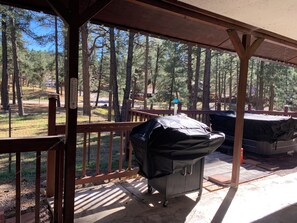 A covered outdoor area with a grill and barbecue equipment, surrounded by a wooden deck and a view of a forested area.