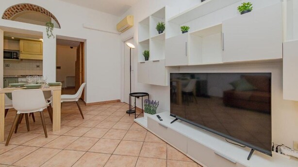 Uma sala de estar moderna com uma televisão de tela grande, uma área de jantar com mesa e cadeiras, e estantes embutidas com plantas.