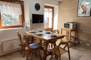 A dining area with a wooden table set for four, surrounded by wooden chairs with patterned cushions. A television is mounted on the wall above the table, and a poster is visible on the adjacent wall.