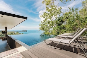 Une piscine à débordement avec des chaises longues, une terrasse en bois et une vue sur la mer et les îles.
