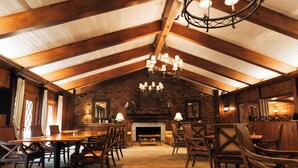 A spacious dining area with a high ceiling and exposed wooden beams, featuring a central chandelier and a brick fireplace. - Pine Needles Lodge & Golf Club (Southern Pines)