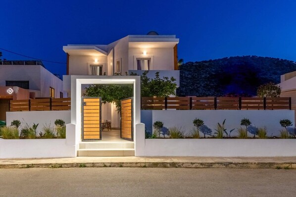 A modern two-story house with a well-lit entrance, a wooden fence, and a landscaped garden. - Villa Suntouch by Villa Plus (Malia)