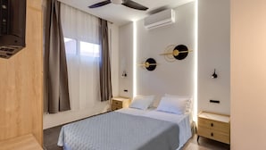 Quarto com cama, mesas de cabeceira de madeira, ventilador de teto, aparelho de ar condicionado e janela com cortinas.