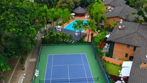 Un court de tennis entouré d’une végétation luxuriante et bordé d’une piscine, avec vue sur l’océan et une montagne à l’horizon.