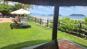 Une vue sur la plage depuis un espace abrité, équipé de chaises longues et surmonté d’un toit de chaume.