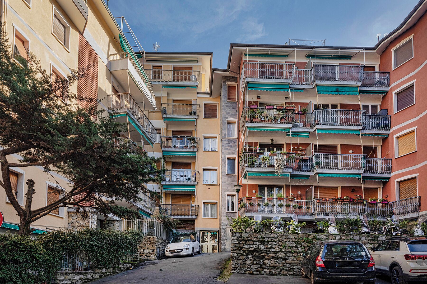A multi-story residential building with balconies, a stone wall, and parked cars.