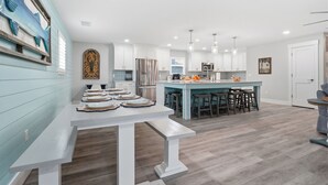 A modern kitchen with white cabinetry, a central island with stools, and a dining table with plates. - 8BR Luxe Retreat w/ Pool, Spa & Rooftop Deck, Beach + Bay Views, Walk to Beach (Bradenton Beach)