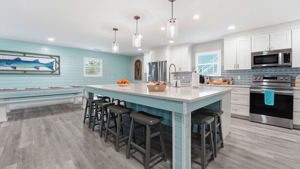 A modern kitchen with a central island, bar stools, stainless steel appliances, and a light blue accent wall. - 8BR Luxe Retreat w/ Pool, Spa & Rooftop Deck, Beach + Bay Views, Walk to Beach (Bradenton Beach)