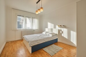 Une chambre à coucher avec un lit, une fenêtre avec des stores, un radiateur et un luminaire suspendu.