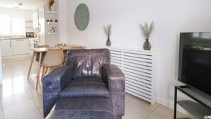 Una sala de estar moderna con un sofá de cuero, un área de comedor con mesa y sillas, una cocina al fondo, un reloj en la pared y un televisor sobre un mueble.
