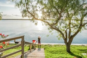 A wooden deck leading to a lake with a person fishing, surrounded by greenery and a tree. - Relaxed 3BR Near the Beach | Pool + Dock Access (Corpus Christi)