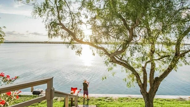 A wooden deck leading to a lake with a person fishing, surrounded by greenery and a tree. - Near the Beach 3BR Stay | Poolside BBQ + Fishing (Corpus Christi)