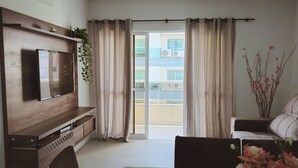 Un salon avec un téléviseur fixé à un mur en bois, une table à manger recouverte d’une nappe à motifs floraux et un canapé.