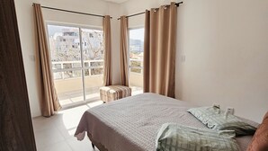 Une chambre avec un grand lit, un petit tabouret et une porte-fenêtre coulissante donnant sur un balcon.