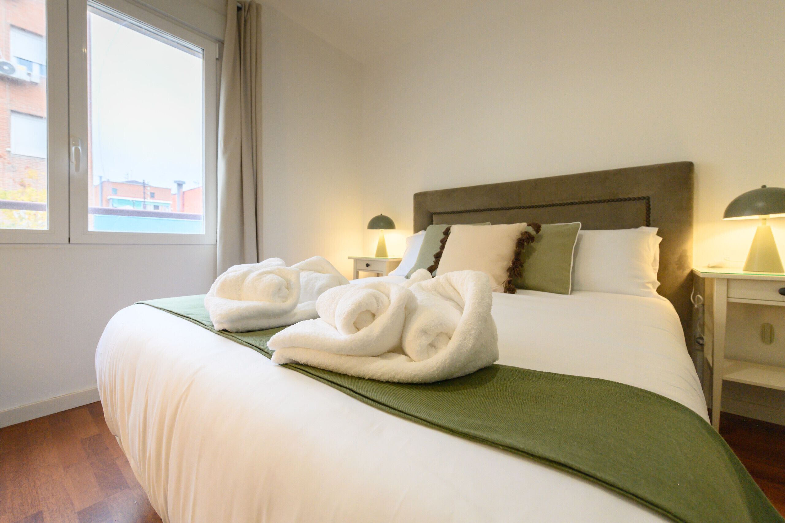 Una cama bien hecha con sábanas blancas y detalles verdes, dos lámparas de noche y vistas de edificios a través de una ventana.