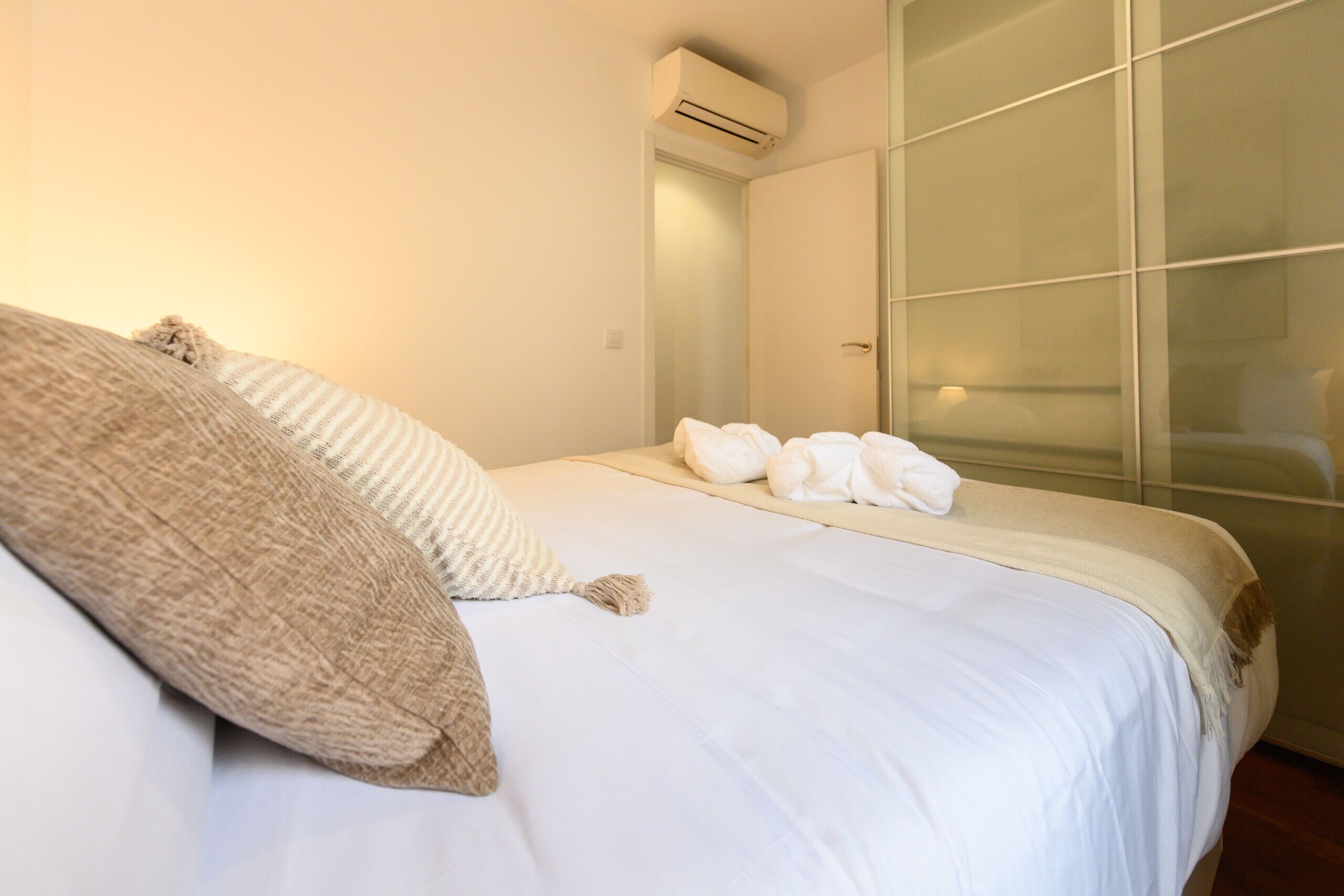 Een net opgemaakt bed met witte lakens en een beige plaid, een glazen scheidingswand en een aan de muur bevestigde airconditioning.