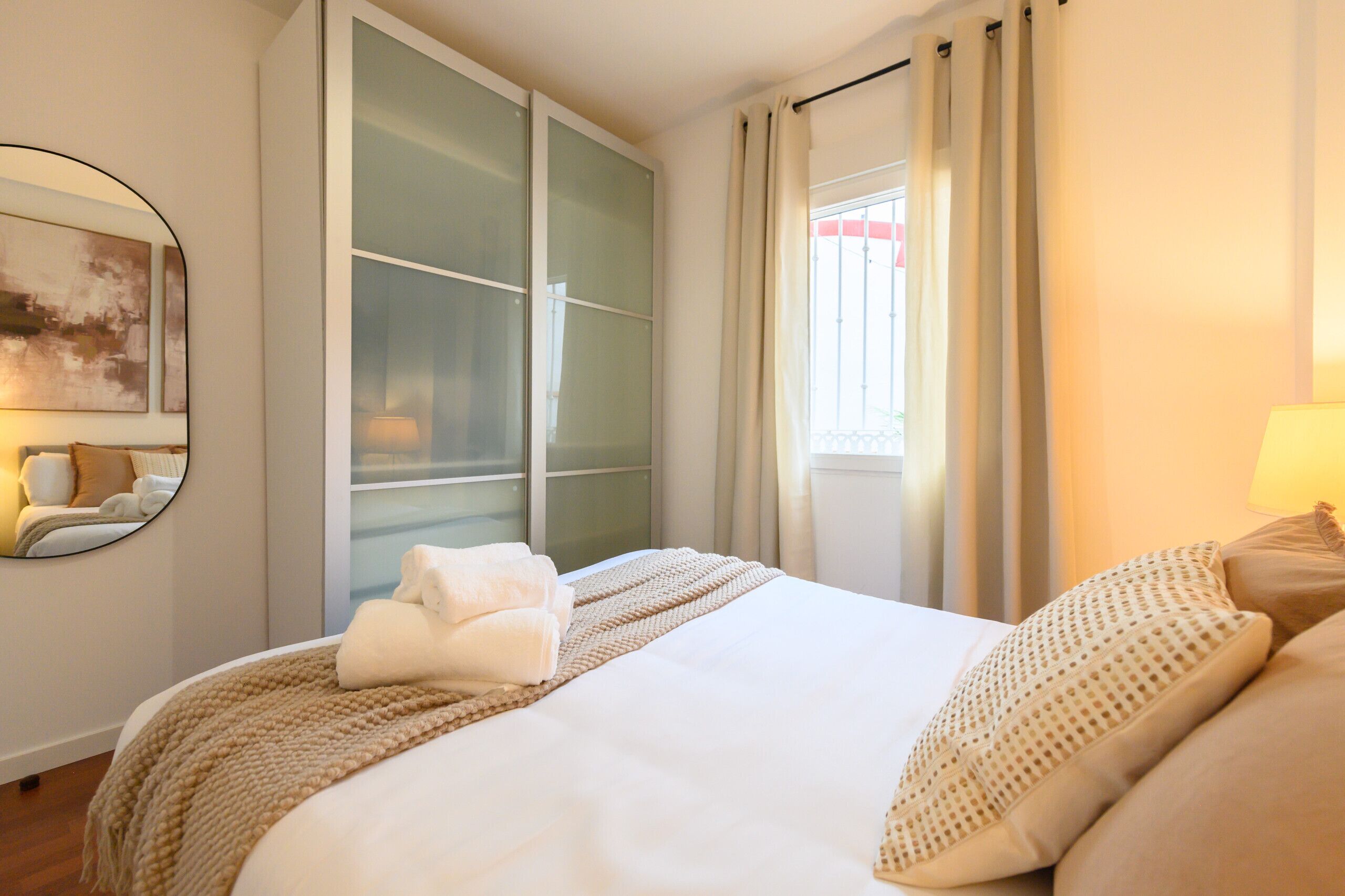 A hotel room with a bed, a large window with curtains, a wardrobe, and a mirror on the wall.