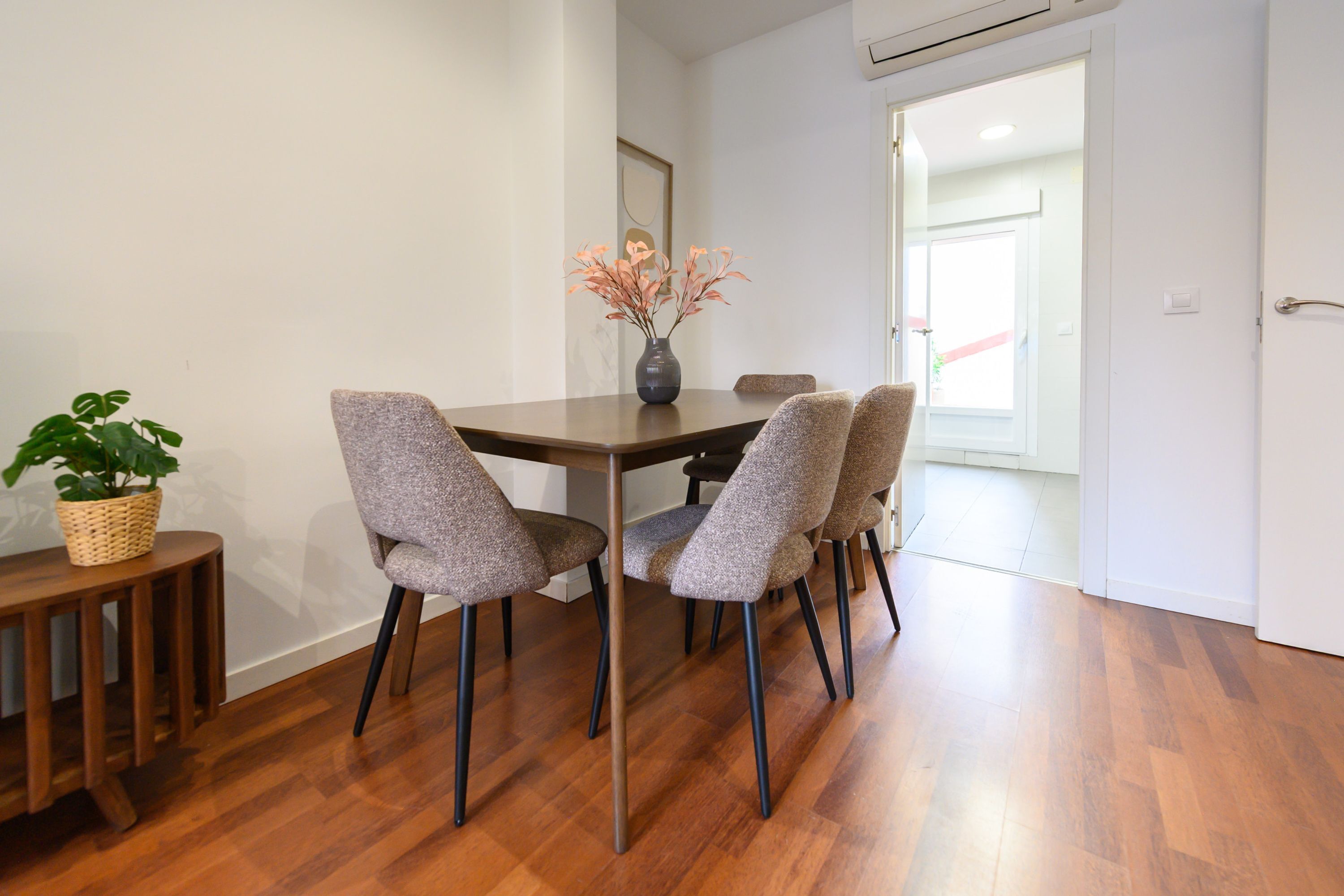 A dining area with a wooden table, four grey upholstered chairs, a vase with flowers, and a wicker basket with a plant.
