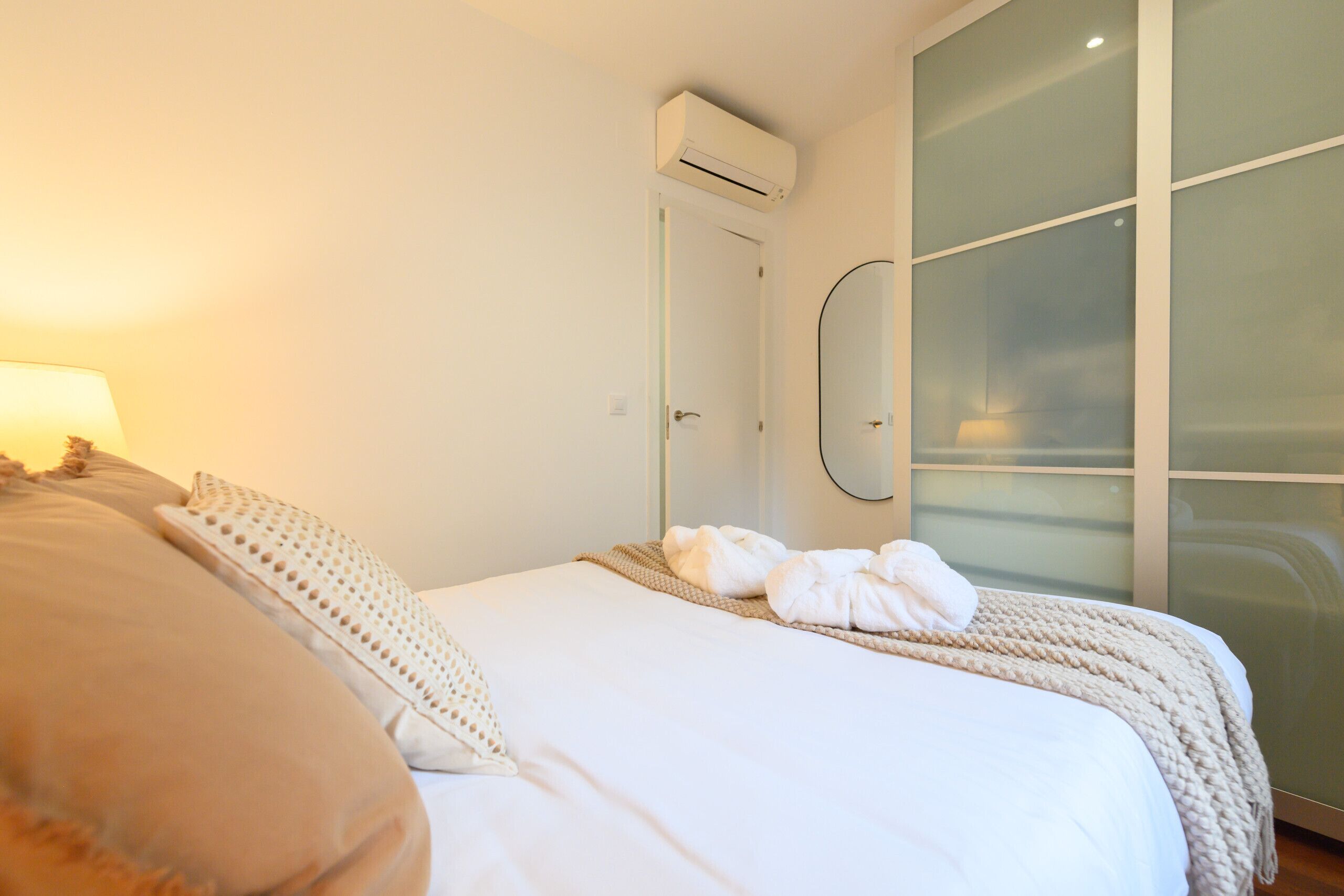 A hotel room with a bed, a wall-mounted air conditioner, a glass partition, and a mirror.