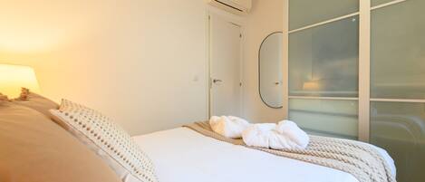 A hotel room with a bed, a wall-mounted air conditioner, a glass partition, and a mirror.