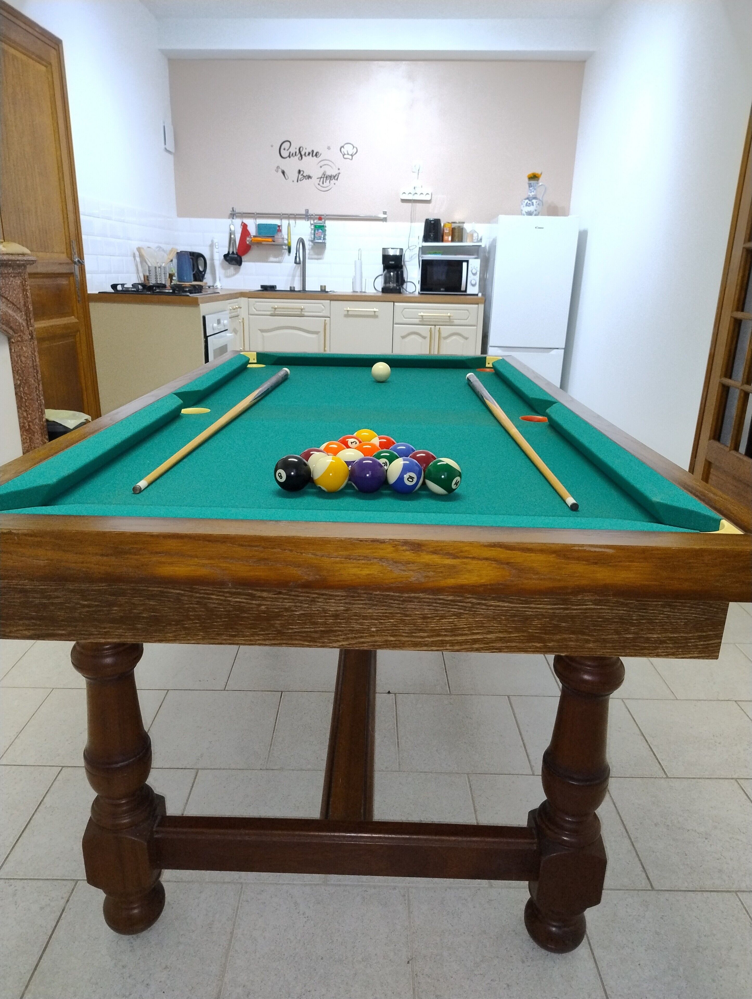 A pool table with balls, a kitchen area with appliances, and a wall with a sign.