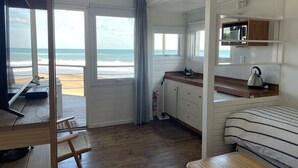 Une chambre située en bord de mer, avec un plancher en bois, un lit avec une literie à rayures, une kitchenette équipée d’une bouilloire et une vue sur l’océan.