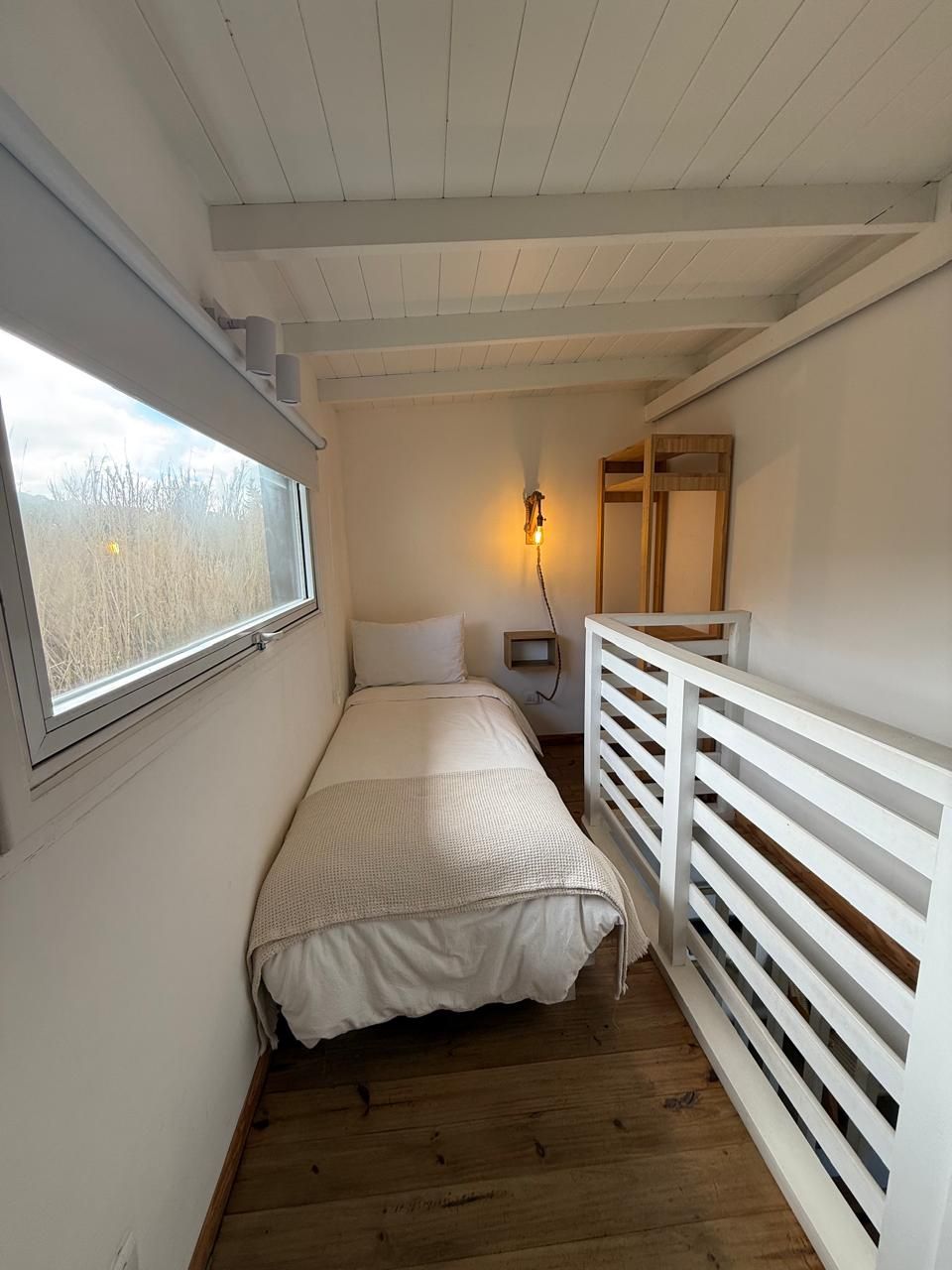 A bedroom with a bed, a nightstand, a bedside lamp, a window, and a wooden floor.