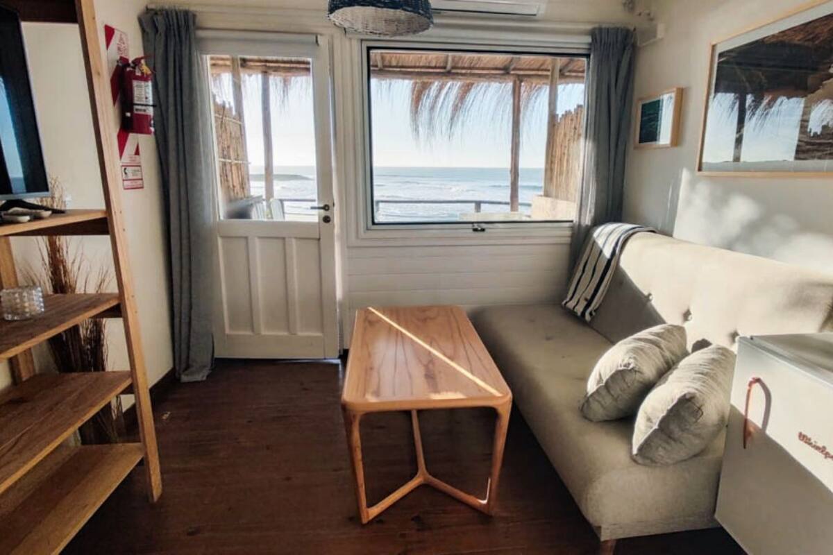 Una habitación con una mesa de madera, un sofá con almohadones, un televisor y una ventana con vistas al mar.