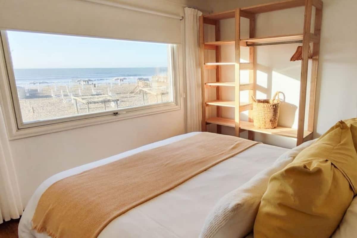 Un dormitorio con cama, una ventana con vista al mar, una repisa de madera con una cesta y una lámpara.