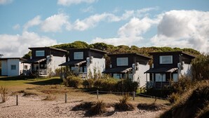 Une rangée de petits chalets situés au bord de la plage, avec une plage de sable au premier plan, une zone herbeuse et une colline boisée en arrière-plan.